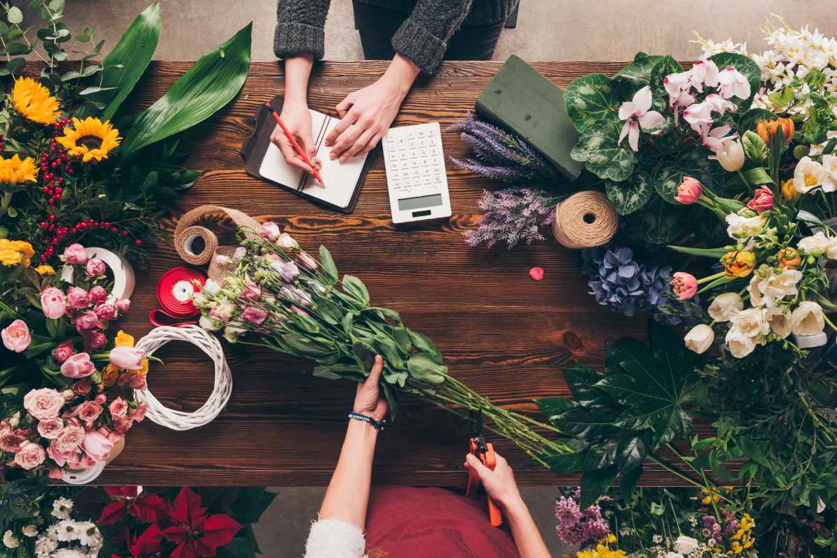 Zwei Personen arrangieren und planen Blumenarrangements auf einem Holztisch mit verschiedenen Blumen und Floristikwerkzeugen.
